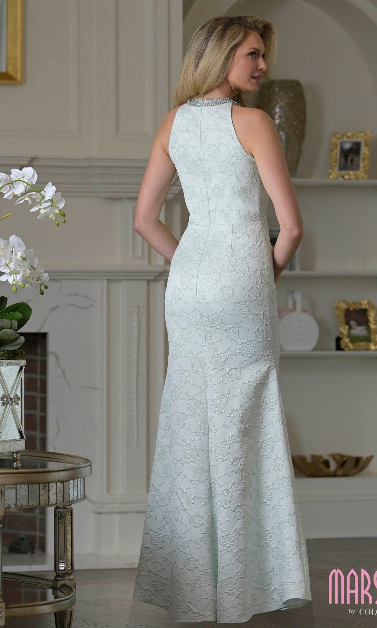 A woman stands indoors facing away, wearing the Marsoni M356 long formal dress in light blue floral jacquard with a high neckline.